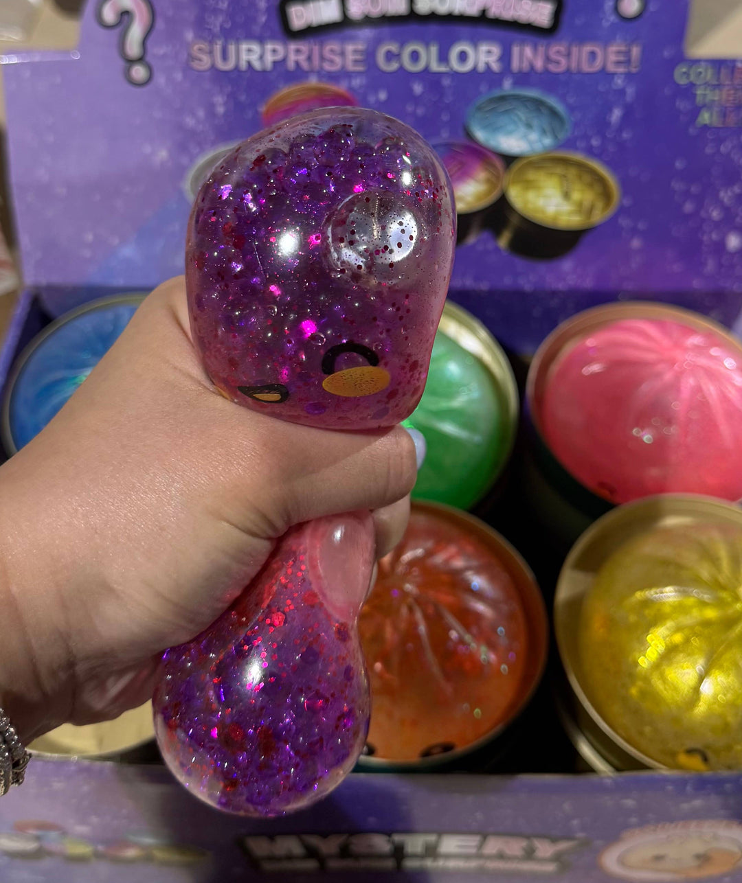 A hand holds a purple glitter surprise toy inside a cardboard display box filled with colorful surprise toys. The display card reads 'Mystery One Size Surprise' and shows multiple round surprise capsules in different colors.