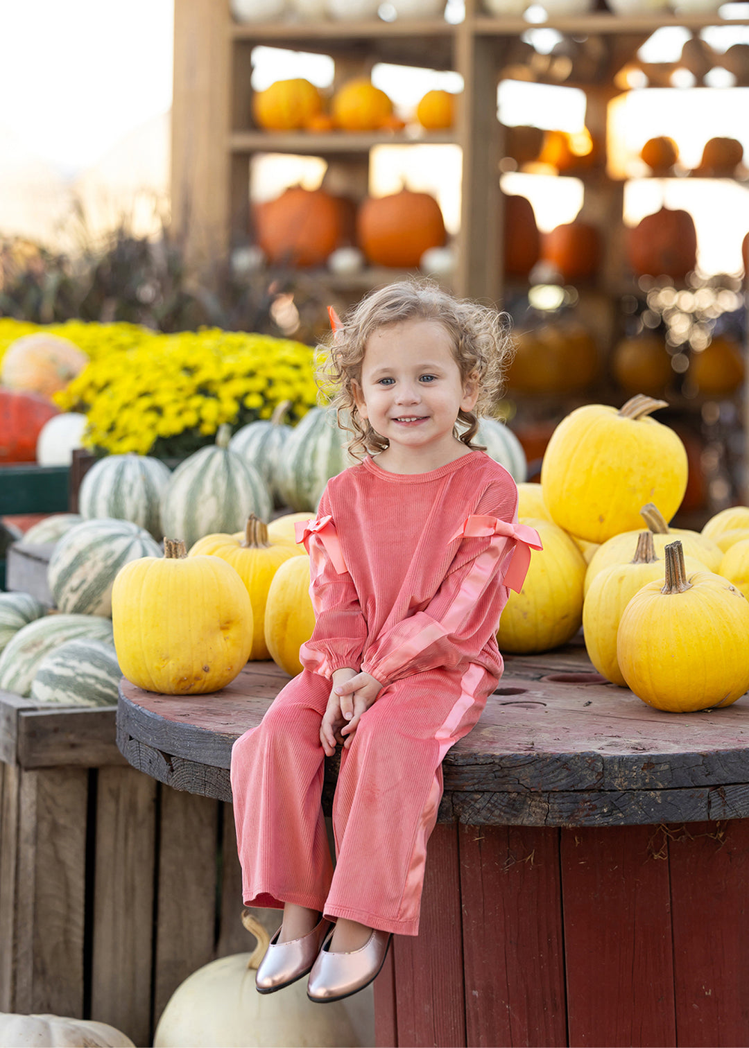 A young girl with curly blonde hair sits on a wooden barrel at a pumpkin patch, wearing a matching dusty pink long-sleeve jumpsuit and metallic silver shoes. Yellow pumpkins, striped gourds, and yellow flowers are visible in the background.