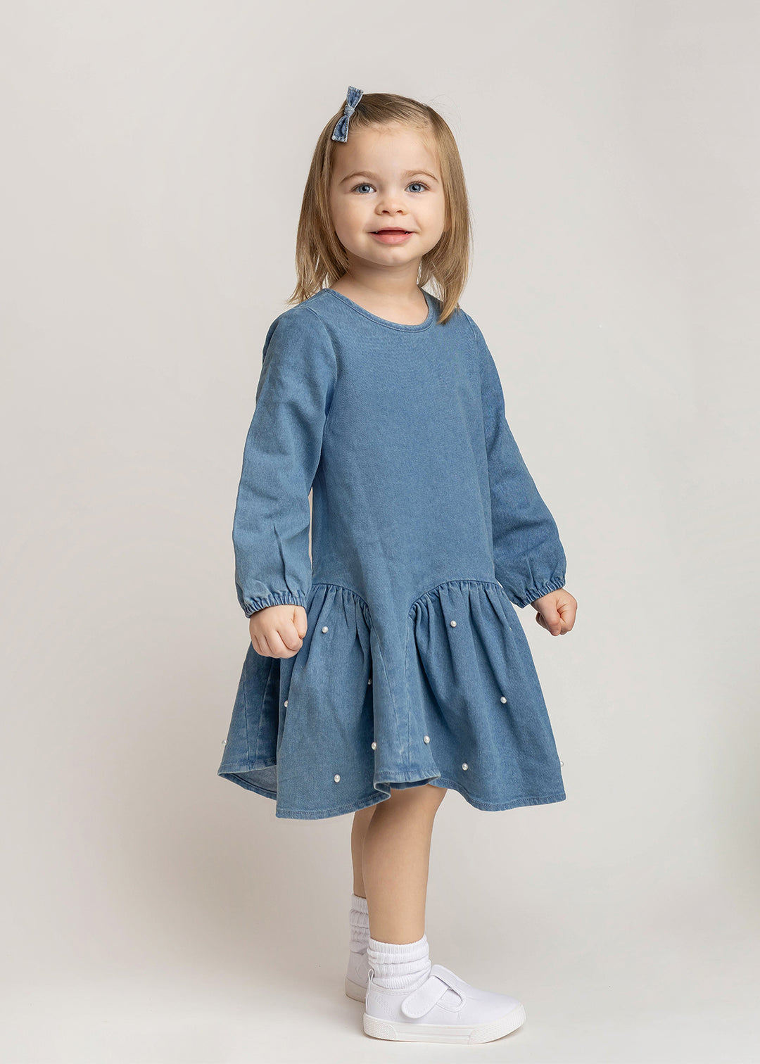 A young girl with blonde hair wearing a dusty blue long-sleeve dress with white embroidered pearls, white socks, and white sneakers, standing against a light beige studio background.