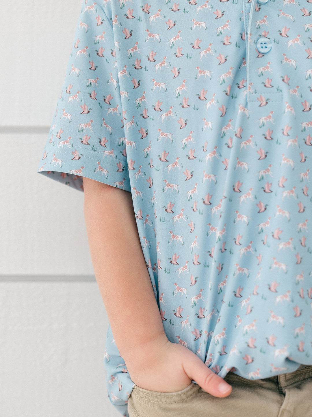 A close-up of a child wearing a light blue short-sleeve polo shirt with a repeating pink and white unicorn print, paired with khaki pants.