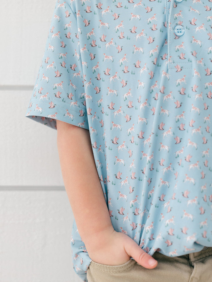 A close-up of a child wearing a light blue short-sleeve polo shirt with a repeating pink and white unicorn print, paired with khaki pants.