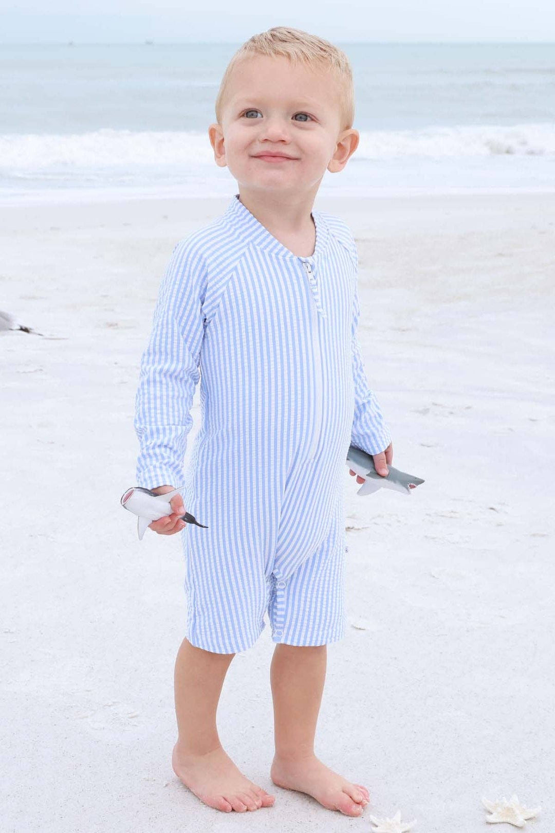 Child in a blue striped swimsuit standing on a sandy beach.