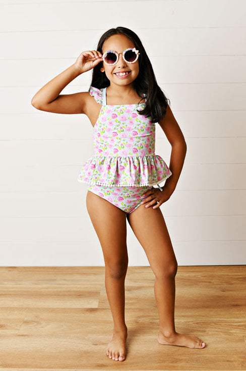 Young girl wearing a floral swimsuit and sunglasses on a wooden floor with a white background