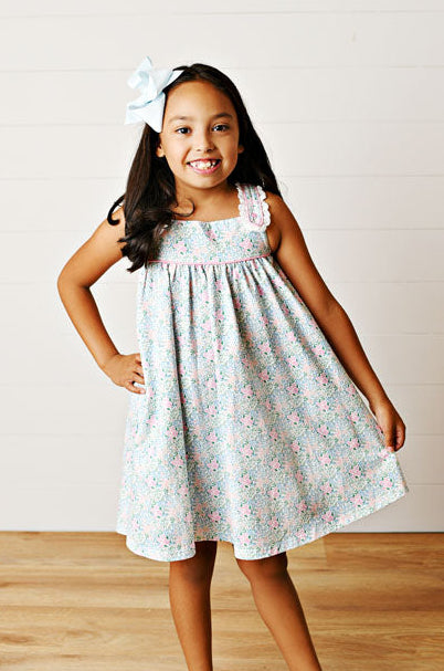 Young girl wearing a light blue floral dress with white sandals on a wooden floor.