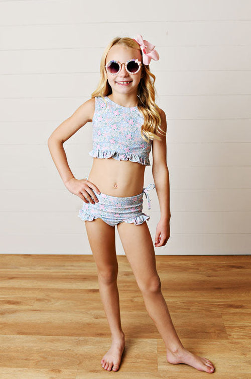 Young girl in a colorful two-piece swimsuit with sunglasses and a flower in her hair on a wooden floor.