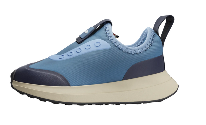 A light blue and navy slip-on sneaker with a thick cream-colored midsole, shown against a dimmed gray background with other shoe options partially visible.