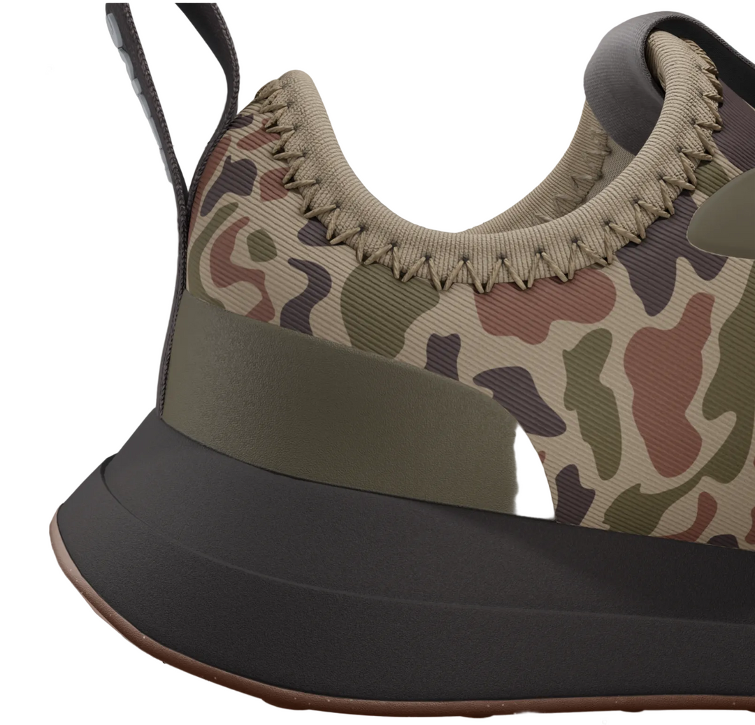 A close-up of a sneaker shown on a product page, featuring a beige, green, and brown camouflage pattern on the upper, a thick olive green midsole band, and a dark gray outsole.