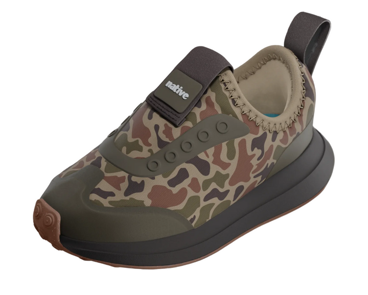 A single slip-on water shoe with a green, brown, and beige camouflage pattern, dark gray straps and sole, and a tan rubber toe cap, shown against a faded product page background.