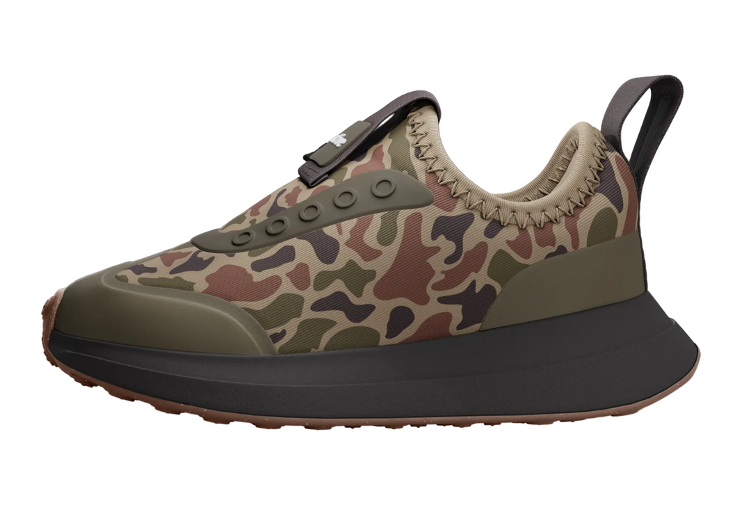 A low-top sneaker with a camouflage pattern upper in beige, brown, and green, featuring an olive green lace guard, beige stitching, a black pull tab, and a thick black and brown sole, shown on a dim gray background.