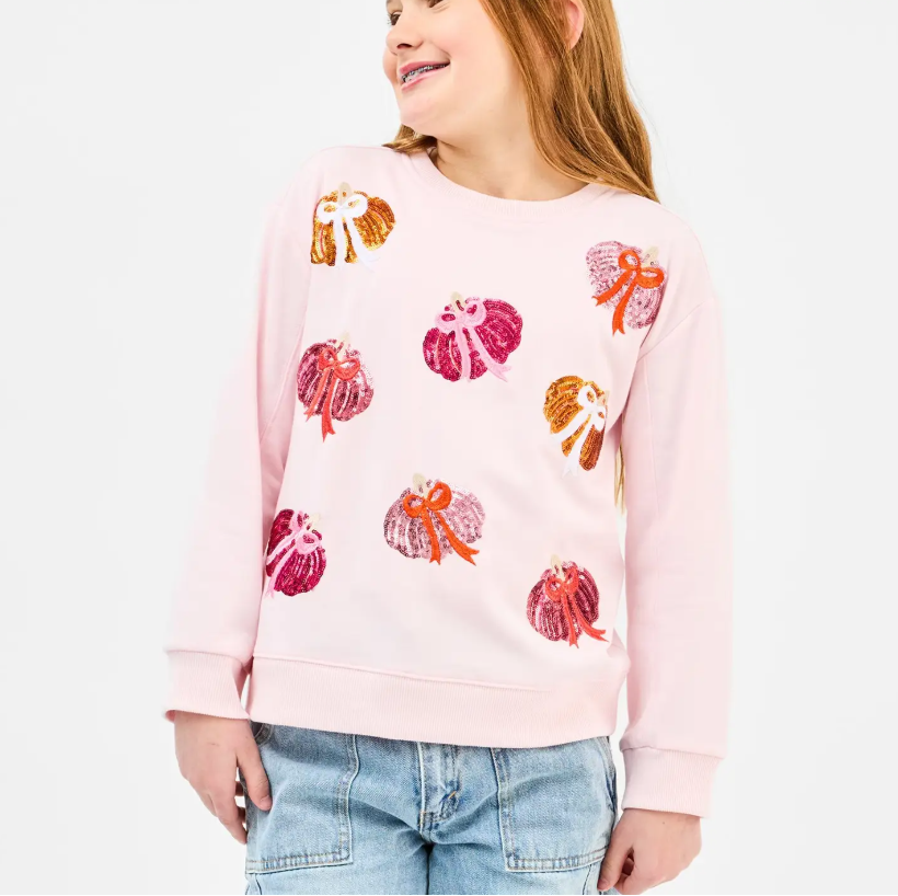 A young girl wearing a light pink crewneck sweatshirt decorated with colorful embroidered pumpkins and blue jeans, shown against a plain white background.
