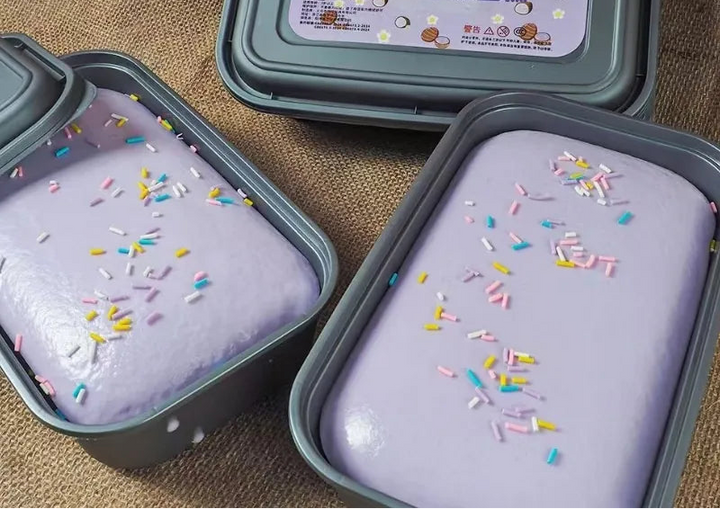 Two containers of purple cake with colorful sprinkles on a textured surface