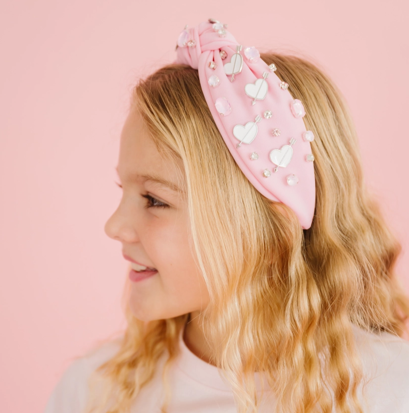 Young girl wearing a pink headband with heart decorations against a pink background