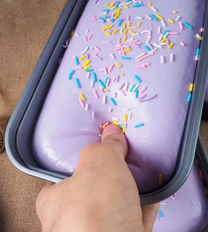 Purple ice cream with colorful sprinkles in a container, touched by a hand.
