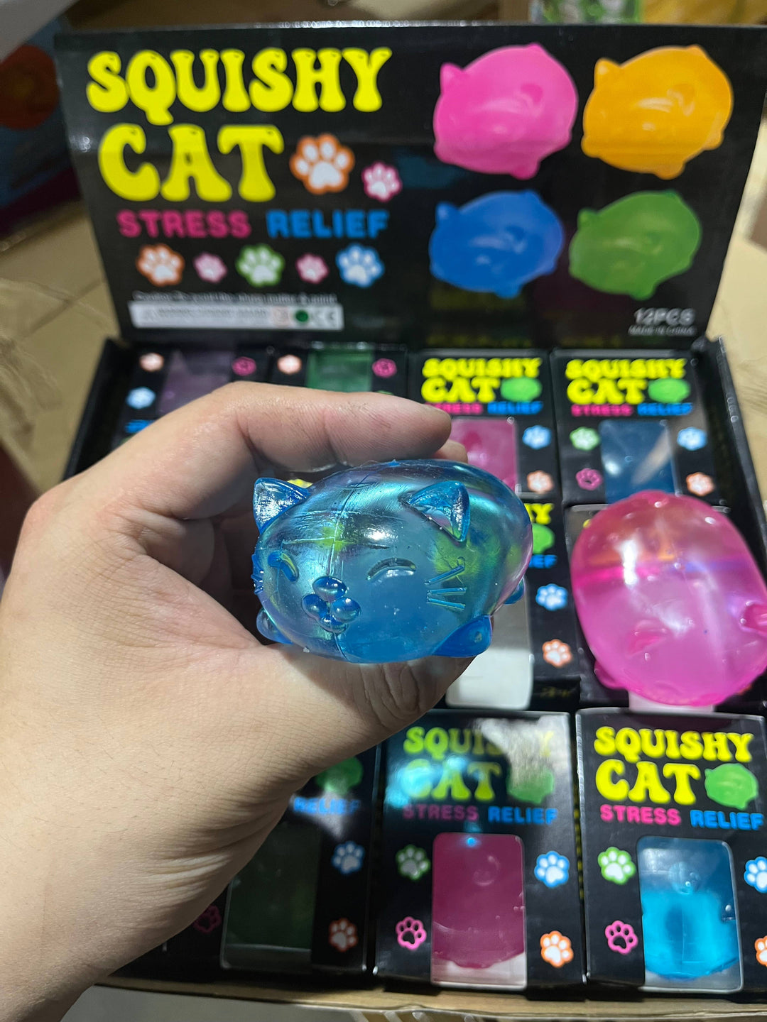 Blue squishy cat toy held in front of a box labeled 'Squishy Cat Stress Relief' with colorful toys.